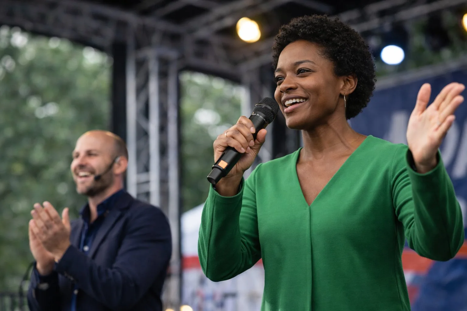 Une femme noire souriante en robe verte parle dans un microphone, un homme applaudit en arrière-plan.